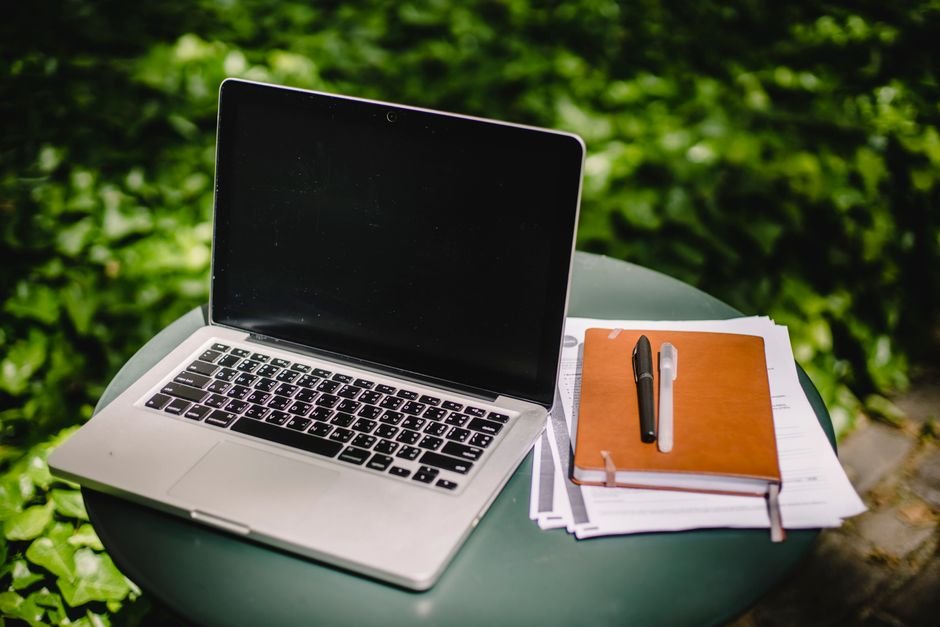 Modern netbook with empty screen placed near brown notebook and papers on round table against blurred green garden