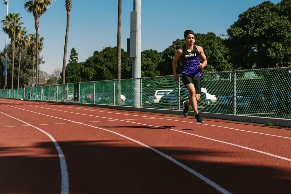 A young male athlete in a purple jersey running on an outdoor track with palm trees.