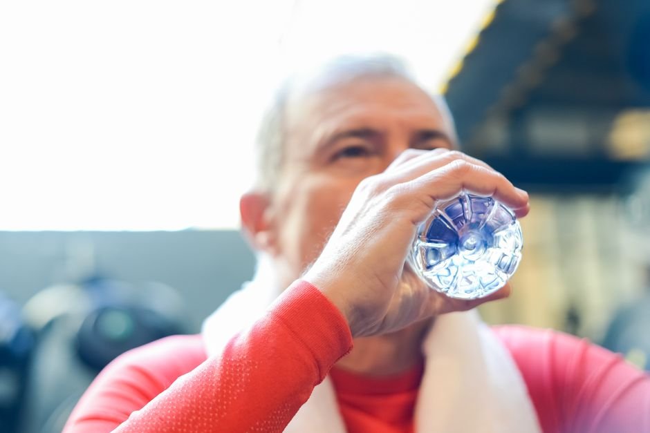 Elderly man in red shirt sipping water during gym session.