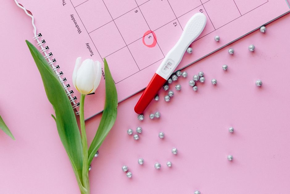 A close-up of a pregnancy test, calendar, and tulip arranged on a pink surface.