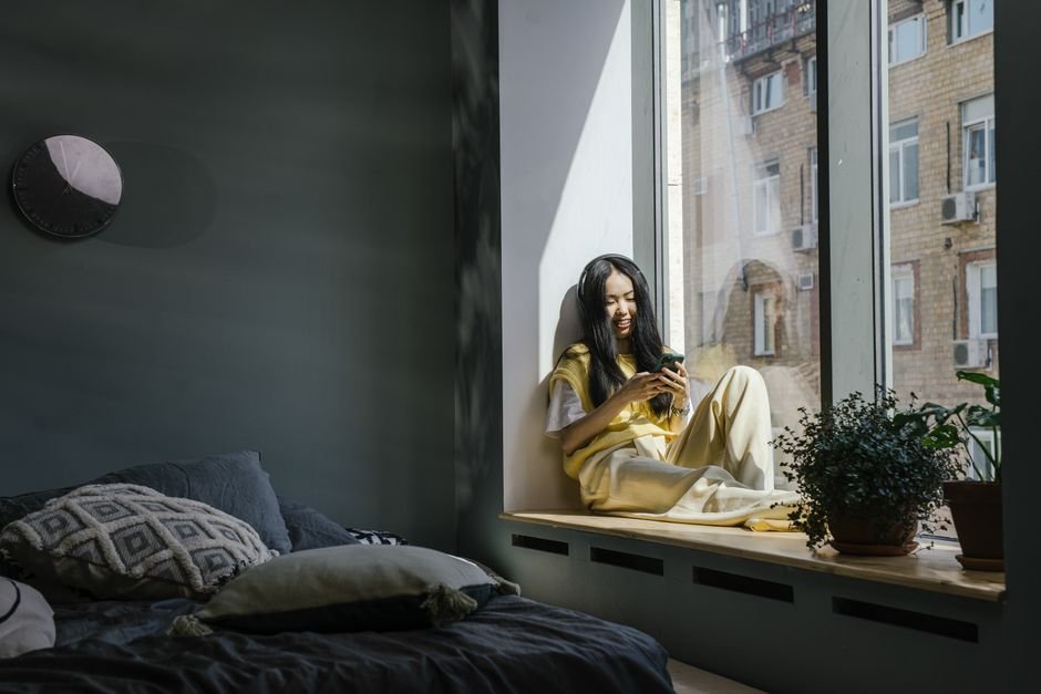 Asian woman sitting by a window indoors, smiling at her phone in a cozy home setting.