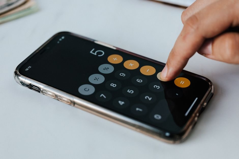Crop faceless financier touching mobile phone screen during work while calculating on calculator application representing digits and financial symbols