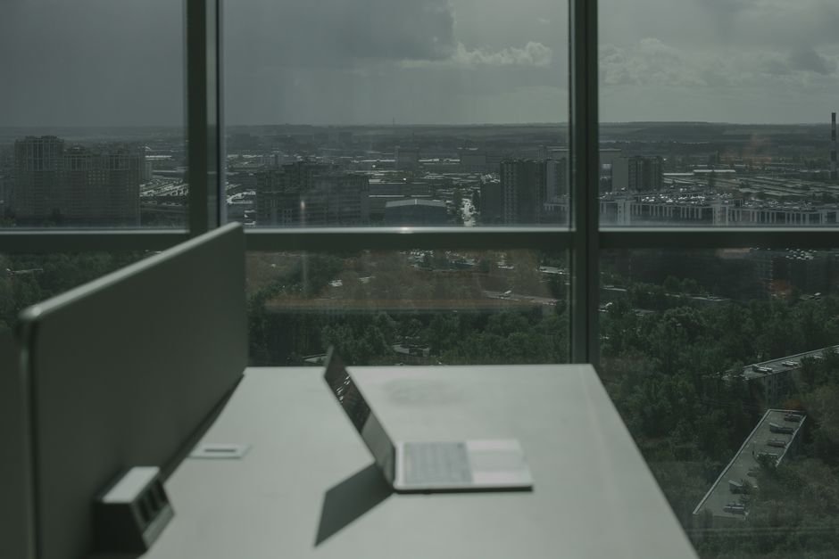 Laptop on office desk with a city skyline through a large window, offering a modern workspace view.