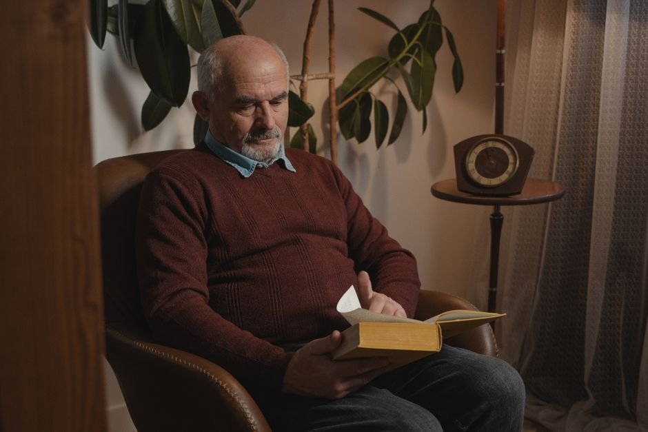 A senior man with facial hair reading a book indoors, surrounded by plants and a clock.