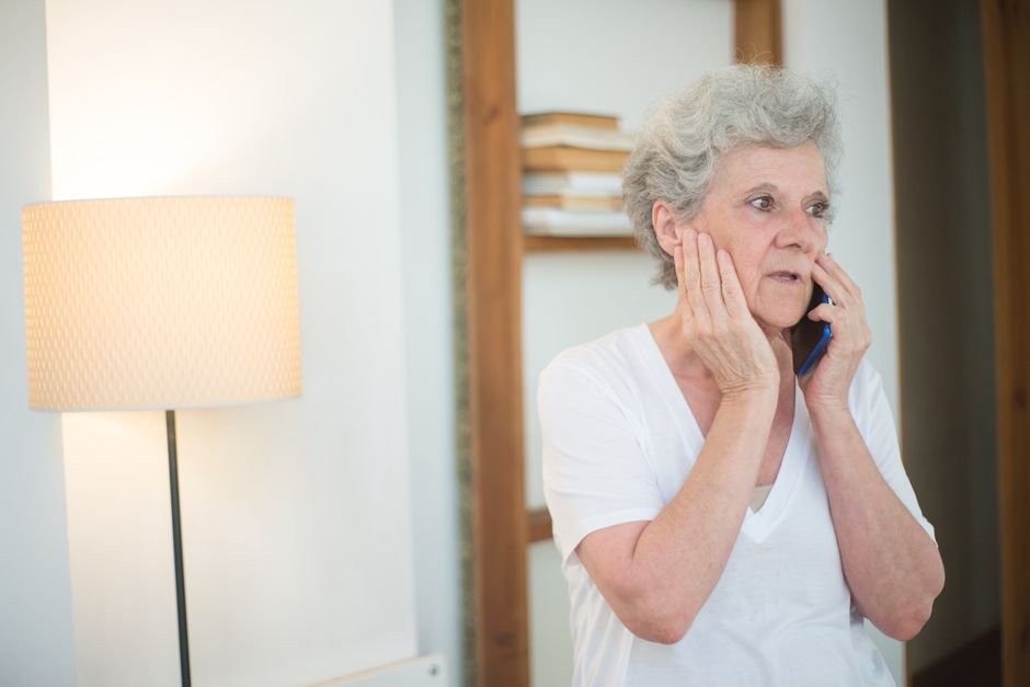Elderly woman with white hair talking on phone, appearing concerned, indoors.
