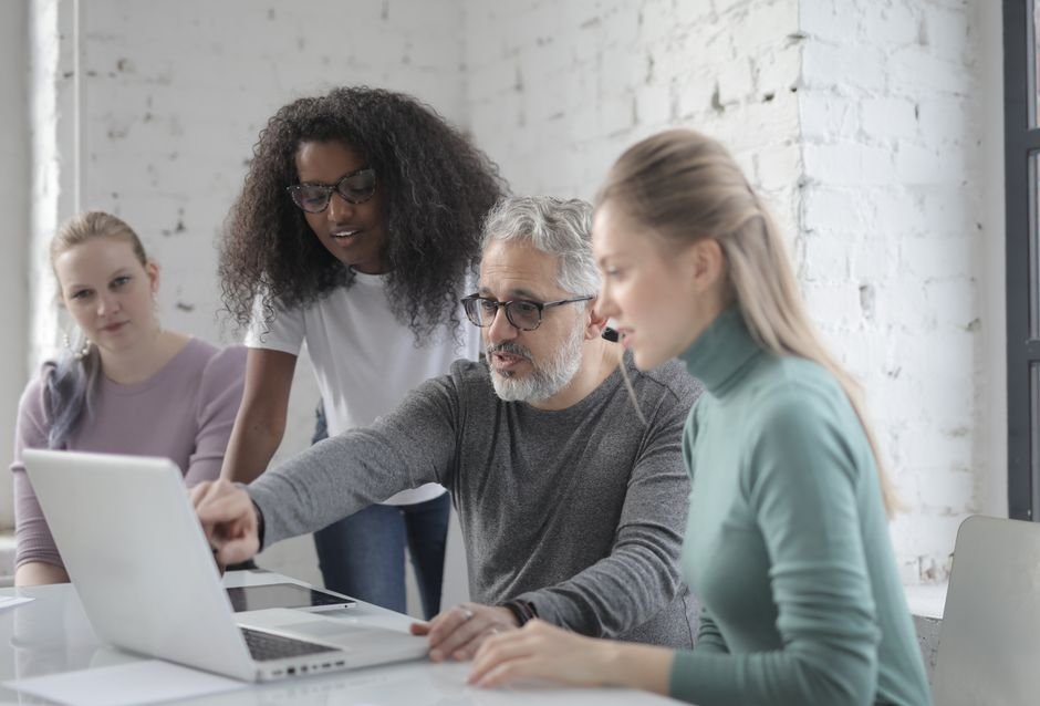 Bearded man wearing casual clothes sitting at table and working on laptop and explaining details of project to women while working