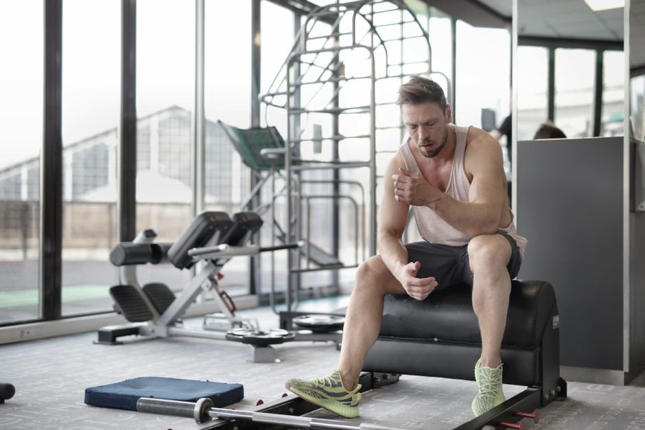 Tired young muscular male athlete in casual sportswear and sneakers sitting on bench and resting after intense weightlifting workout in modern fitness club