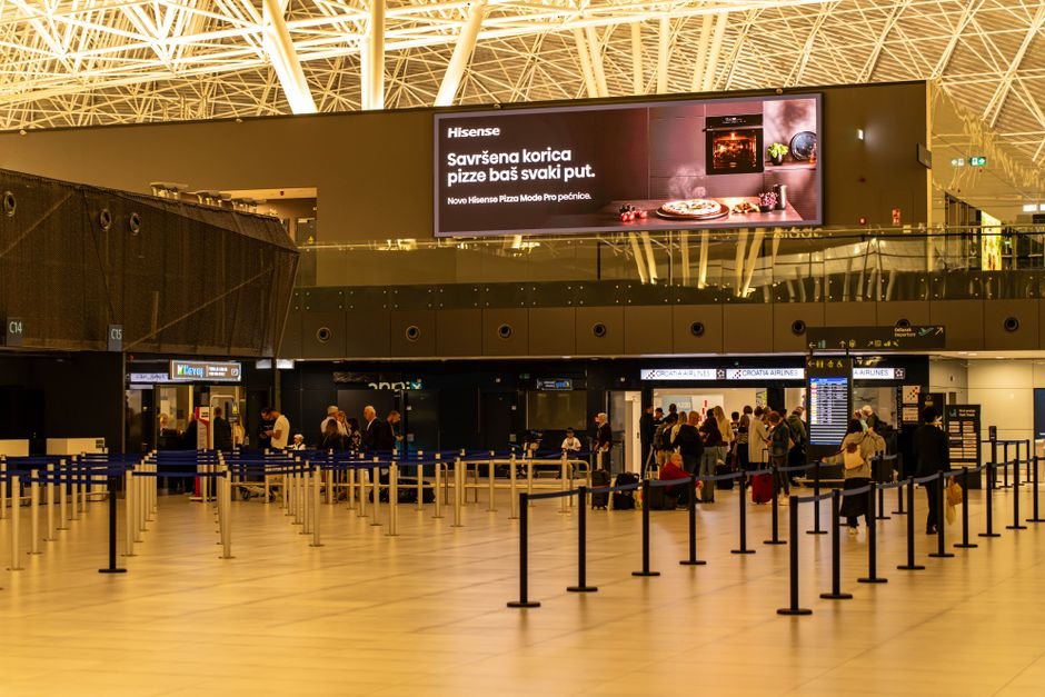 Travelers check in at Zagreb Airport with large advertising screen above.