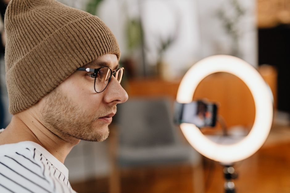 A man wearing eyeglasses and a knitted cap records a video at home using a smartphone and ring light.