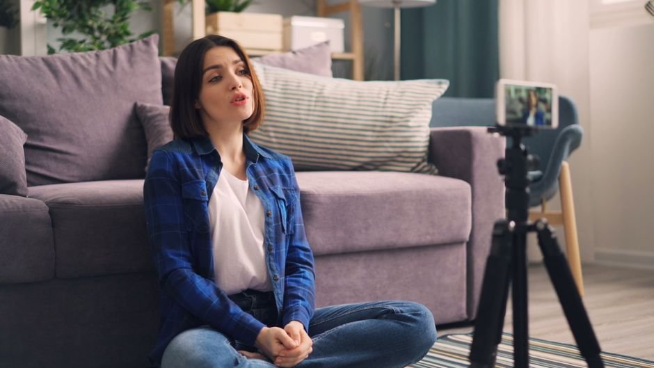 Adult woman recording video indoors on a smartphone, sitting cross-legged on the floor.