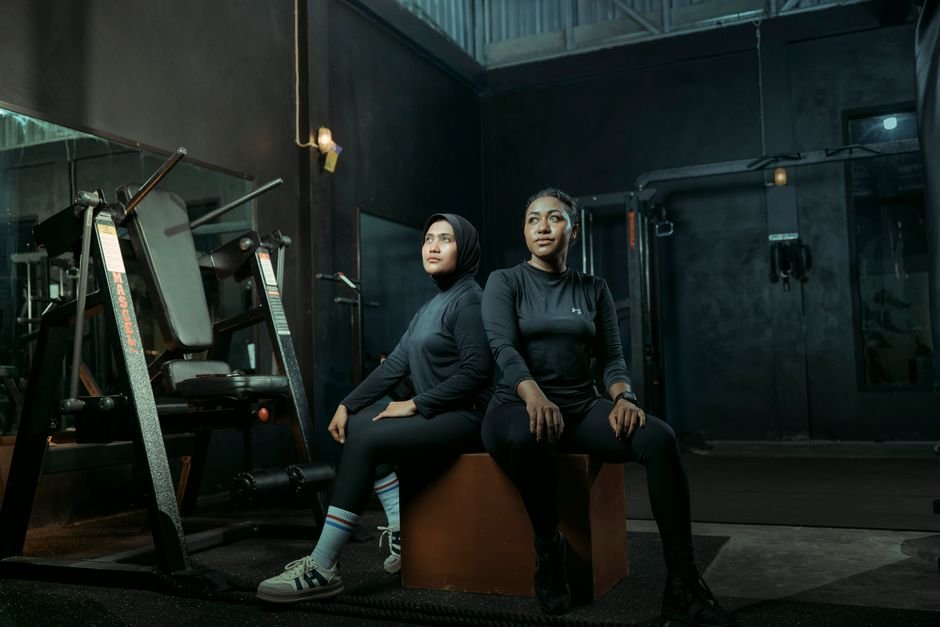 Two women in athletic wear sitting on a box with gym equipment in the background.
