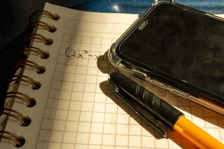A notebook with a math equation, smartphone, and pen on checkered paper in warm lighting.