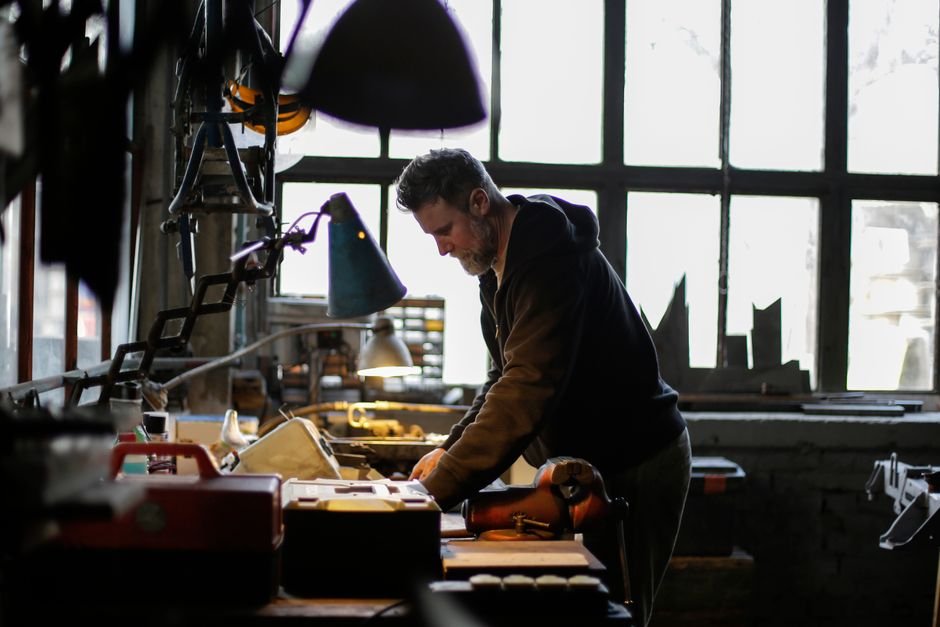 Side view of serious adult bearded male mechanic in workwear standing near workbench and working with details of engine in light of lamp in garage