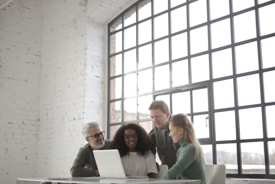 Positive focused multiracial coworkers gathering together near table with laptop in workplace at industrial building during remote work on team against big window at daytime